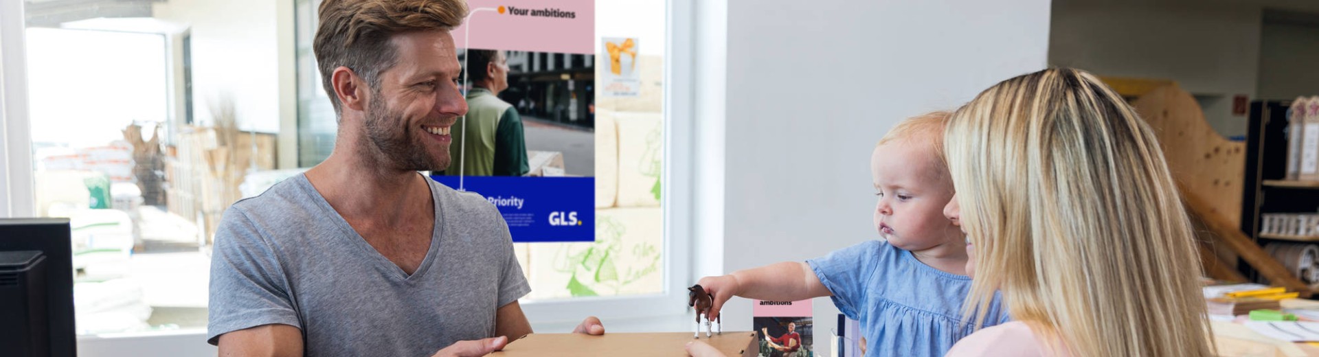 A woman holding her daughter hands a package to a man working at a parcel shop.